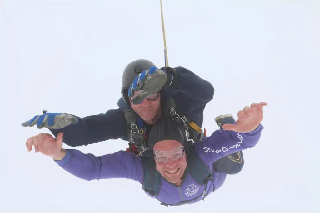 Skydiver in mid-air with parachute cords attached, against a clear sky background.
