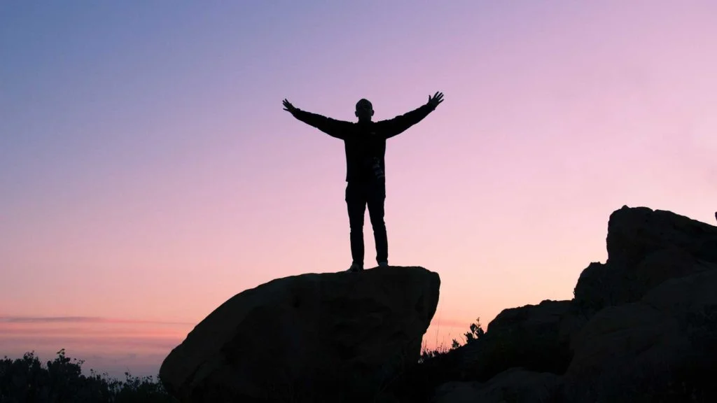 Silhouette of a person with arms raised standing on a rock against a twilight sky.