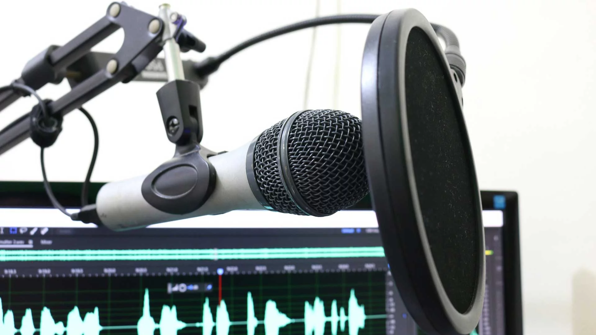 Microphone with pop filter mounted on an arm stand, with waveform on computer screen in background.
