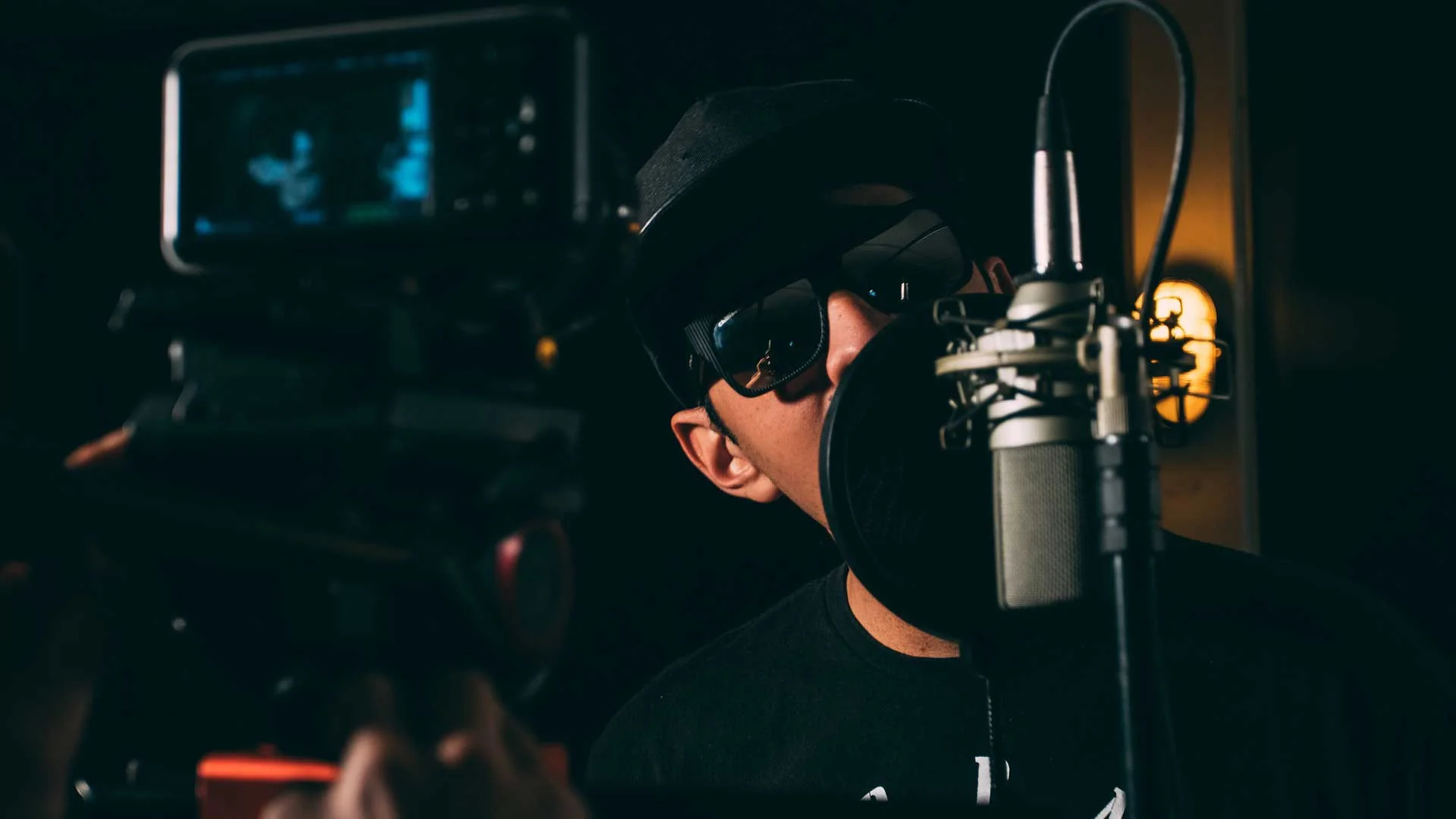 Person in a hat recording at a microphone with camera and ambient lighting in the background.