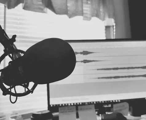 A black and white photo of a microphone in a studio with a blurred computer monitor in the background.