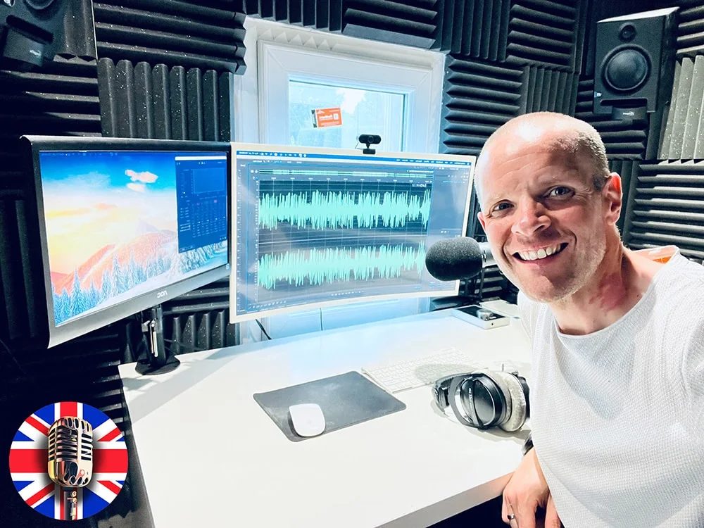 Affordable Voice Over Artist, Neil Williams, in his professional home recording studio. He is sat to the right of the image, smiling at the camera. To the left of him is his microphone along with two computer monitors and a white desk.