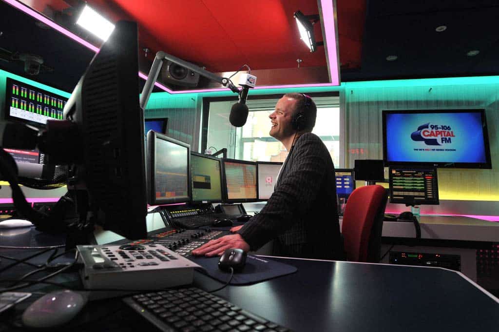 London Voice Over, Neil Williams in the Capital FM studio. Its a side on image of him talking into a microphone, sat at a mixing desk, surrounded by computer monitors, faders and knobs.
