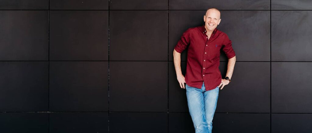 Image of professional voice over from London, Neil Williams. He is leaning against a dark wall, to the right of the image. He is wearing a burgundy ralph lauren shirt, with blue jeans, his legs crossed and hands on his hips.