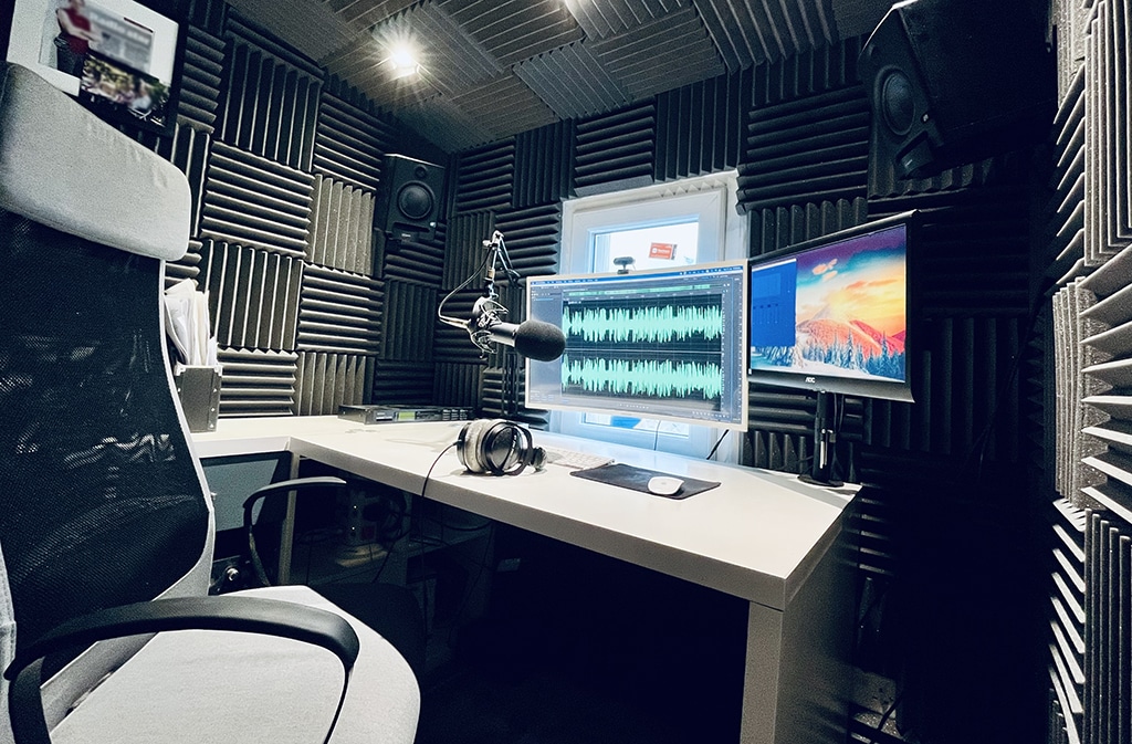 The recording studio of neil williams who offers cheap voice over rates by having his own studio. The image shows an office chair to the left of the screen, with a white desk in front of it. Above that are two computer monitors, one large on the left and a smaller one on the right. There is a microphone in front of them. There are soundproofing panels on the walls, grey in colour.