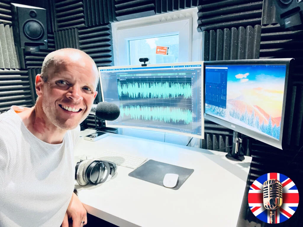 An image of Neil Williams, voice talent online, in his home recording studio in London, UK, where he voices his recordings. In the image you can see Neil on the left, sat in front of the microphone he uses to record the english voiceover artist samples you can hear. There are two computer monitors in the image.