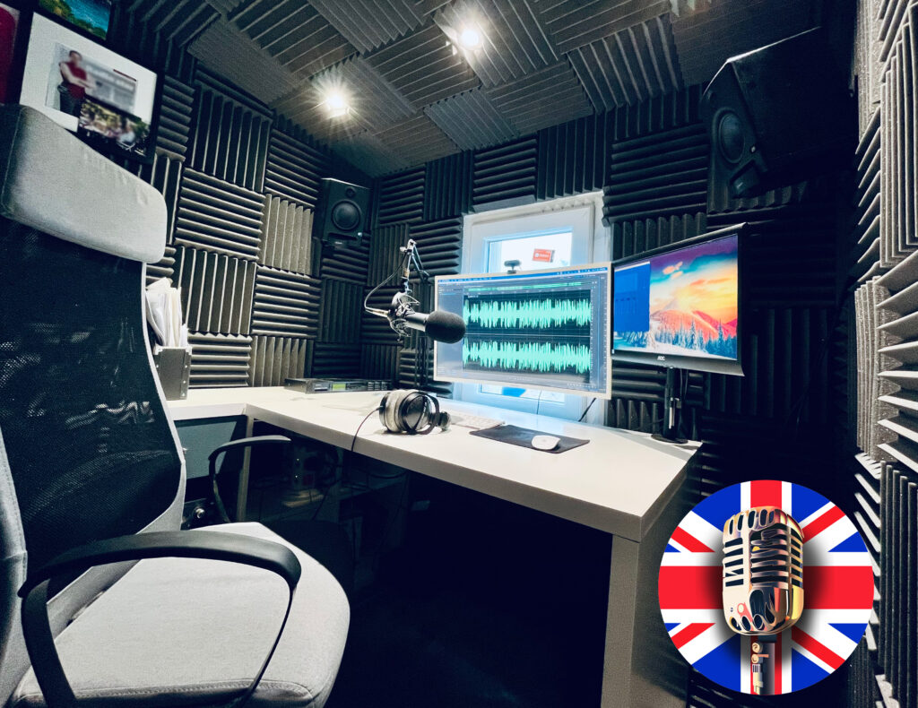 studio of professional voice over in London, UK, Neil Williams. There is a large office chair to the left of the image with a white desk in front. Above the desk are two computer monitors, one is displaying an audio file being edited in adobe audition, the other has a standar wallpaper. There is a microphone on a microphone arm, along with studio monitor speakers and sound proofing on the wall and ceiling, which is grey in colour.