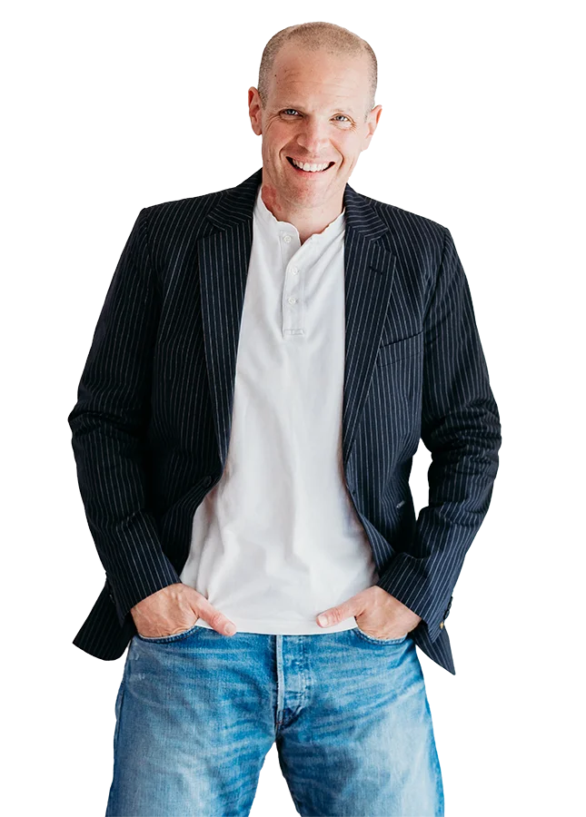 Neil Williams available to hire voice over talent online. He is wearing blue jeans, a white tshirt and black pin striped jacket, standing with his hands in his jean pockets and smiling.